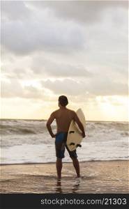 Rear view of young man surfer with white surfboard on a beach at sunset or sunrise
