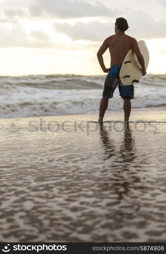 Rear view of young man surfer with white surfboard on a beach at sunset or sunrise