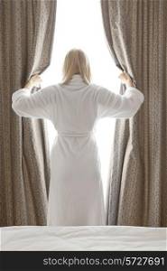 Rear view of young woman in bathrobe opening bedroom curtains at hotel room