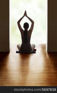 Rear view of young woman meditating on hardwood floor while raising hands