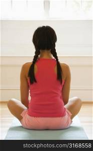 Rear view of young woman sitting on mat with legs crossed in yoga pose.
