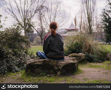 Rear view shot of a young woman sitting on a rock in the park, there is a pagoda in the distance