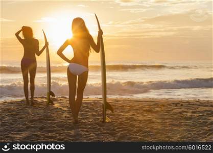 Rear view silhouette of two beautiful sexy young women surfer girls in bikini with surfboards on a beach at sunset or sunrise