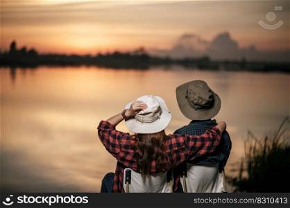 Rear view, Young Asian couple wearing trekking hat sit and enjoy the beautiful nature on sunset near the lake together during c&ing trip