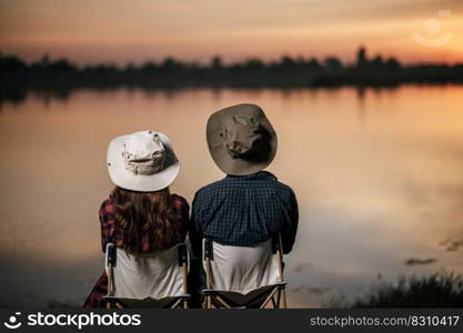 Rear view, Young Asian couple wearing trekking hat sit and enjoy the beautiful nature on sunset near the lake together during c&ing trip