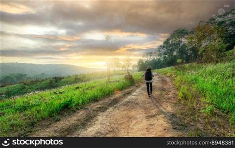 Rearview of relaxed woman walking on road in meadow field to travel in nature with morning sunlight sky. Rural scene. Outdoor activity in summer vacation. Landscape of green grass field and mountains.