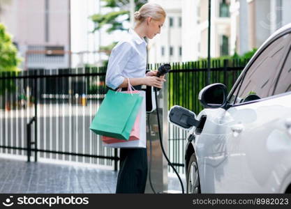 Recharging electric vehicle at charging station with businesswoman carrying shopping bag. Progressive lifestyle of a man in city with ecological concern for clean electric energy driven car ideal.. Recharging EV car with businesswoman carrying shopping bag at charging station.