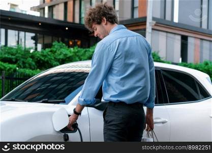 Recharging electric vehicle at charging station with businesswoman carrying shopping bag. Progressive lifestyle of a man in city with ecological concern for clean electric energy driven car ideal.. Recharging EV car with businessman carrying shopping bag at charging station.
