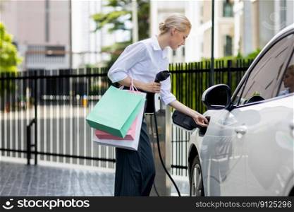 Recharging electric vehicle at charging station with businesswoman carrying shopping bag. Progressive lifestyle of a man in city with ecological concern for clean electric energy driven car ideal.. Recharging EV car with businesswoman carrying shopping bag at charging station.