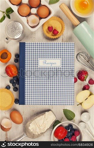 Recipe book, Baking tools and ingredients - flour, rolling pin, eggs, measuring spoons, fruits and berries on vintage wood table. Top view. Rustic background with free text space