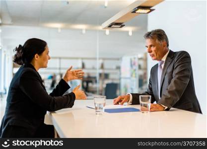 Recruiter (middle aged business man) checking the candidate, an attractive younger woman, during job interview with lively office in the background seen through glass window