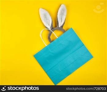 rectangular blue paper shopping bag with a white handle on a yellow background, with bunny ears sticking out of the bag, flat lay