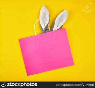 rectangular pink paper shopping bag with a white handle on a yellow background, with bunny ears sticking out of the bag, flat lay