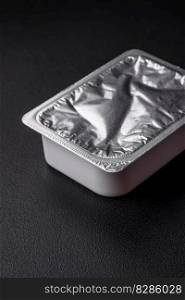 Rectangular plastic box with yogurt or cheese hermetically sealed with a foil lid on a dark concrete background