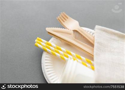 recycling and eco friendly concept - wooden disposable forks, knives, paper cups and straws with canvas napkin on grey background. wooden forks, knives, paper straws and cups