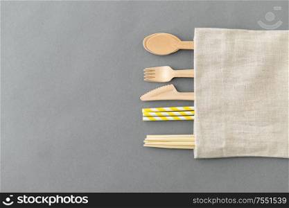 recycling and eco friendly concept - wooden disposable spoons, forks, knives, chopsticks and paper straws with canvas napkin on grey background. wooden spoon, fork, knife, straws and chopsticks