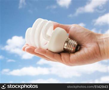recycling, electricity and ecology concept - close up of hand holding energy saving lightbulb or lamp over blue sky and clouds background. close up of hand holding energy saving lightbulb