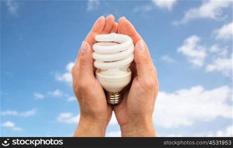recycling, electricity, environment and ecology concept - close up of hands holding energy saving lightbulb or lamp over blue sky and clouds background
