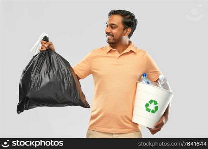 recycling, sorting and sustainability concept - smiling young indian man holding bucket with plastic bottles and trash bag over grey background. smiling indian man sorting paper and plastic waste