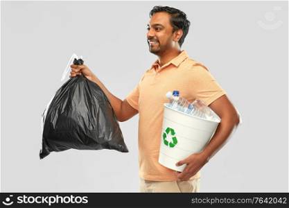 recycling, sorting and sustainability concept - smiling young indian man holding bucket with plastic bottles and trash bag over grey background. smiling indian man sorting paper and plastic waste