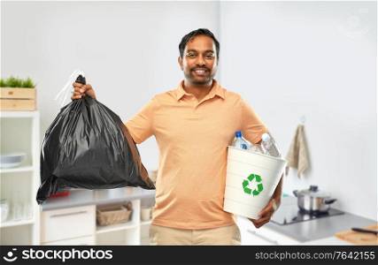 recycling, sorting and sustainability concept - smiling young indian man holding bucket with plastic bottles and trash bag over home kitchen background. smiling indian man sorting paper and plastic waste