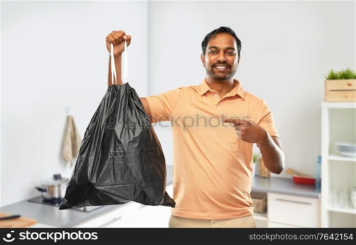 recycling, sorting and sustainability concept - smiling young indian man holding trash bag over home kitchen background. smiling indian man holding trash bag