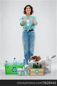 recycling, waste sorting and sustainability concept - happy smiling young woman with plastic and glass bottles, paper and metal tin cans in boxes showing thumbs up over grey background. happy woman sorting paper, metal and plastic waste