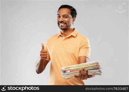 recycling, waste sorting and sustainability concept - smiling young indian man in polo t-shirt with heap of paper magazines showing thumbs up over grey background. indian man with paper waste showing thumbs up