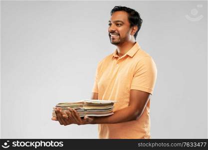 recycling, waste sorting and sustainability concept - smiling young indian man in polo t-shirt holding heap of paper magazines over grey background. smiling young indian man sorting paper waste
