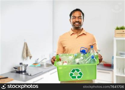 recycling, waste sorting and sustainability concept - smiling young indian man in striped t-shirt holding box with plastic bottles over home kitchen background. smiling young indian man sorting plastic waste
