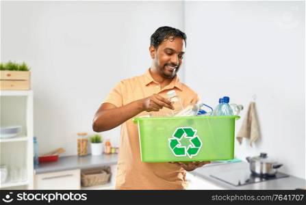 recycling, waste sorting and sustainability concept - smiling young indian man in striped t-shirt holding box with plastic bottles over home kitchen background. smiling young indian man sorting plastic waste
