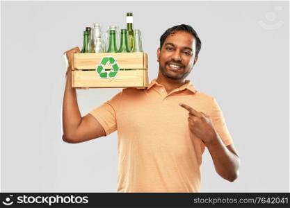 recycling, waste sorting and sustainability concept - smiling young indian man in striped t-shirt holding wooden box with glass bottles and jars over grey background. smiling young indian man sorting glass waste