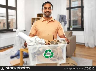 recycling, waste sorting and sustainability concept - smiling young indian man in polo t-shirt holding paper garbage in plastic box over messy home room background. smiling young indian man sorting paper waste