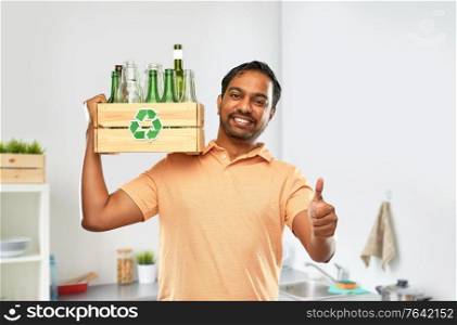 recycling, waste sorting and sustainability concept - smiling young indian man in striped t-shirt holding wooden box with glass bottles and jars and showing thumbs up over home kitchen background. smiling young indian man sorting glass waste