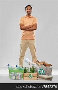 recycling, waste sorting and sustainability concept - smiling young indian man with plastic and glass bottles, papers and metal tin cans in boxes over grey background. smiling man sorting paper, glass and plastic waste