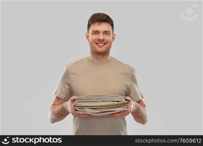 recycling, waste sorting and sustainability concept - smiling young man in striped t-shirt holding heap of paper magazines over grey background. smiling young man sorting paper waste