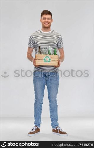 recycling, waste sorting and sustainability concept - smiling young man in striped t-shirt holding wooden box with glass bottles and jars over grey background. smiling young man sorting glass waste