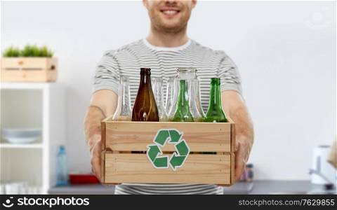 recycling, waste sorting and sustainability concept - smiling young man in striped t-shirt holding wooden box with glass bottles and jars over home kitchen background. smiling young man sorting glass waste