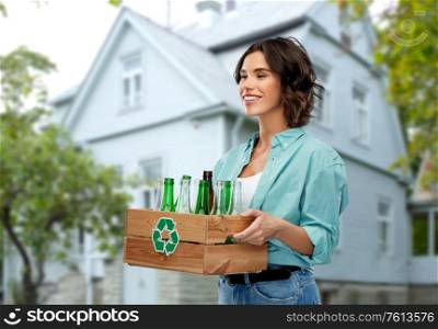 recycling, waste sorting and sustainability concept - smiling young woman holding wooden box with glass bottles and jars over house background. smiling young woman sorting glass waste outdoors