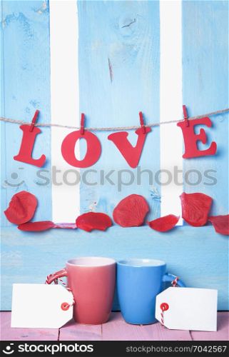 Red and blue cups with blank tags, the word love spelled from red paper letters tied to a string and soap rose petals, on a blue wooden fence