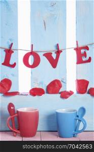 Red and blue mugs with spoons on their handle, the word love written with red paper letters, tied to a string and soap rose petals on a blue fence.