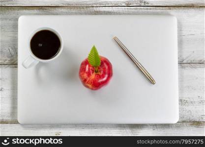 Red apple, coffee and silver pens on top of laptop with rustic white desktop in background.
