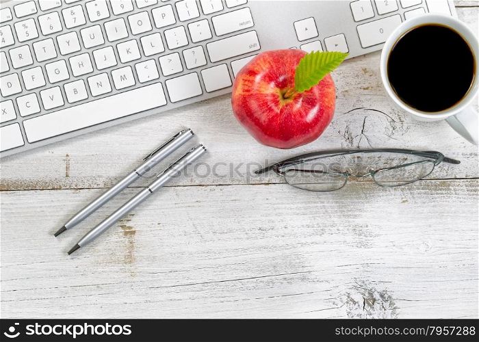 Red apple, coffee, reading glasses and silver pens next to computer keyboard with rustic white desktop underneath.