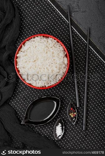 Red bowl with boiled organic basmati jasmine rice with black chopsticks and sweet soy sauce on bamboo placemat with red linen towel on black stone background.