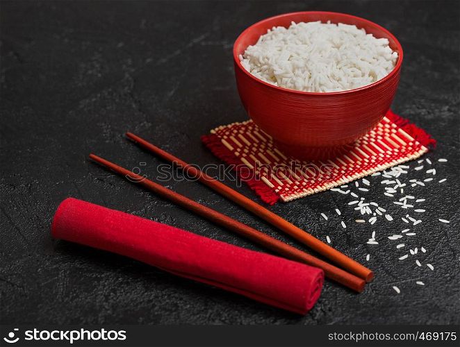 Red bowl with boiled organic basmati jasmine rice with red chopsticks and sweet soy sauce on bamboo placemat with red linen towel on black stone.