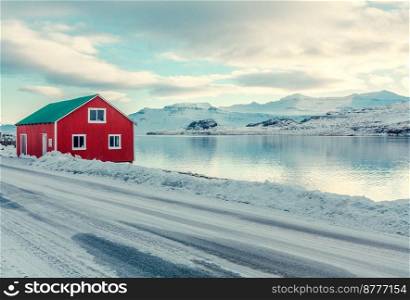 Red cabin on snowy sea coast landscape photo. Beautiful nature scenery photography with mountains on background. Idyllic scene. High quality picture for wallpaper, travel blog, magazine, article. Red cabin on snowy sea coast landscape photo