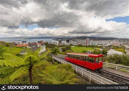 Red cable car moving up the mountain in the foreground and panorama of Wellington in the background. Beautiful blue sky covered with clouds