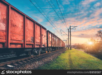 Red cargo wagons and train on railway station against blue sky with clouds at sunset. Colorful industrial landscape with railroad. Railway platform. Heavy industry. Cargo shipping. Transportation. Red cargo wagons and train on railway station