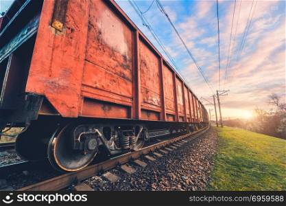 Red cargo wagons and train on railway station against blue sky with clouds at sunset. Colorful industrial landscape with railroad. Railway platform. Heavy industry. Cargo shipping. Transportation. Red cargo wagons and train on railway station
