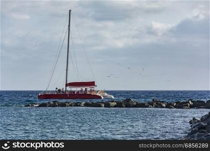 Red catamaran sailing with passengers floating in the sea along the rocks